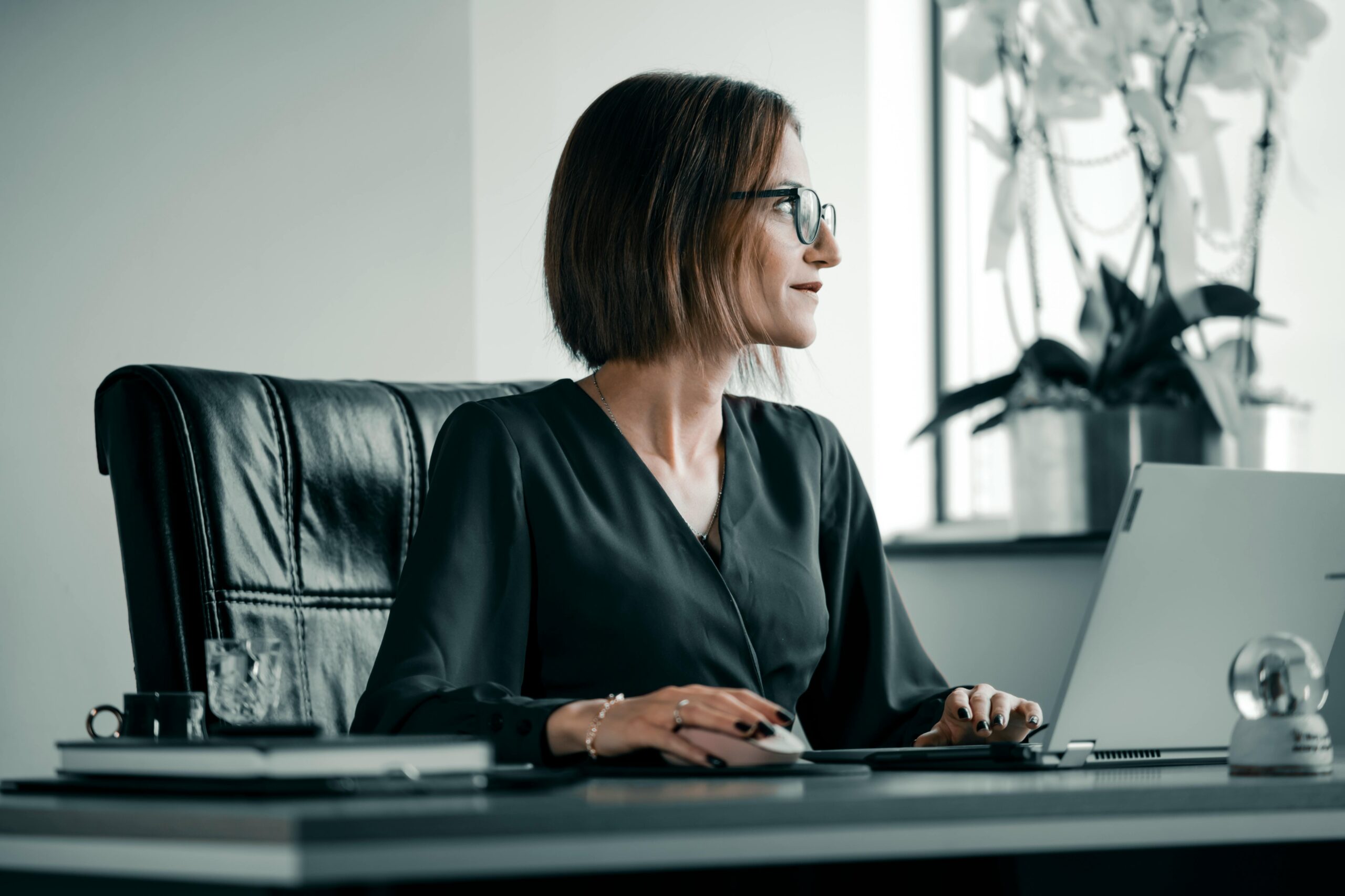 Adult woman working at office desk with laptop, glasses, and leather chair.